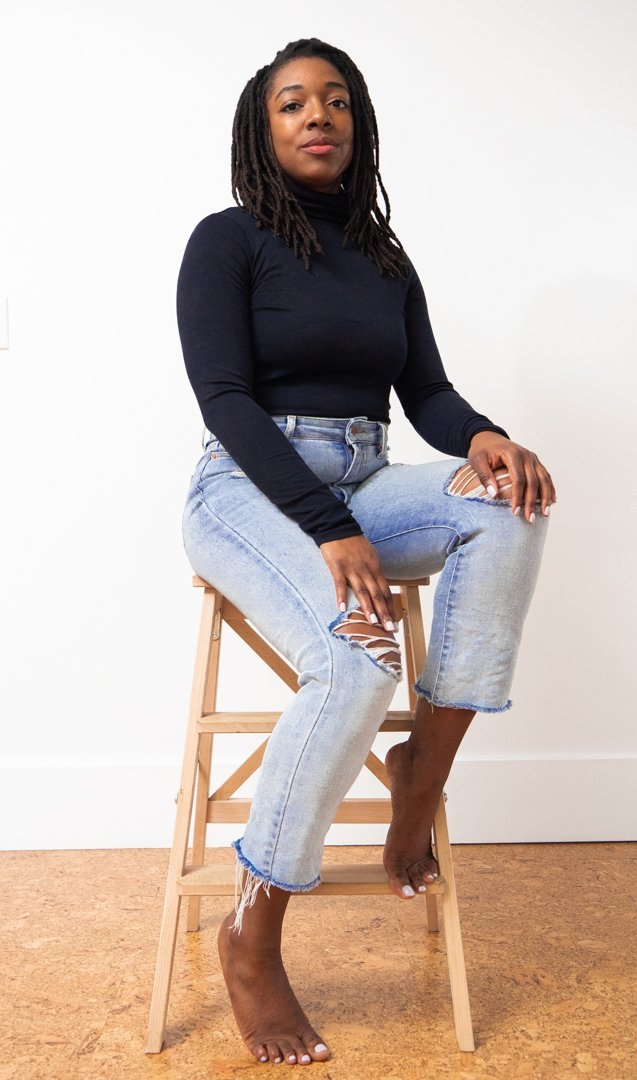 Ana the Marketeer, brand strategist, seated on a stool in a dark turtleneck and light jeans